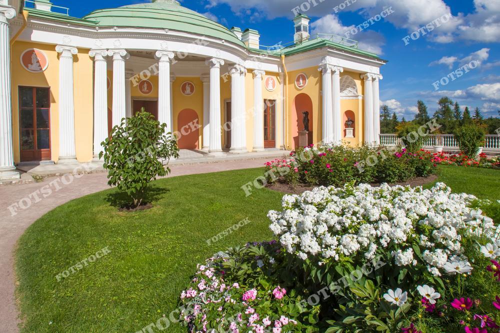 На фотографии изображен павильон в Екатерининском парке, расположенный в Царском Селе. Это архитектурное сооружение в стиле классицизма окружено красивыми цветущими клумбами и зелеными насаждениями. Летнее солнце подчеркивает яркость цветов, а ясное небо создает атмосферу тепла и уюта. Павильон является частью большого комплекса архитектурных и парковых ансамблей, которые входят в состав музея-заповедника.